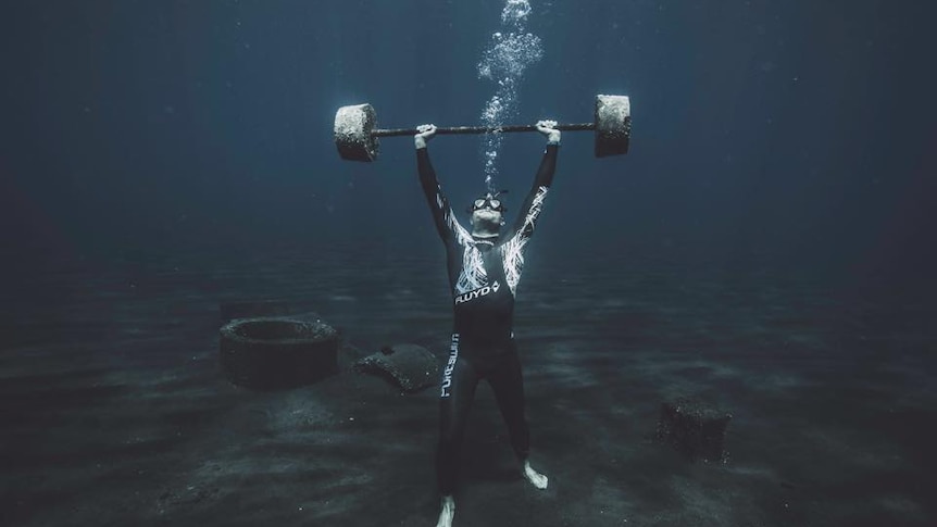 A man standing on the bottom of the ocean holding gym weights above his head.