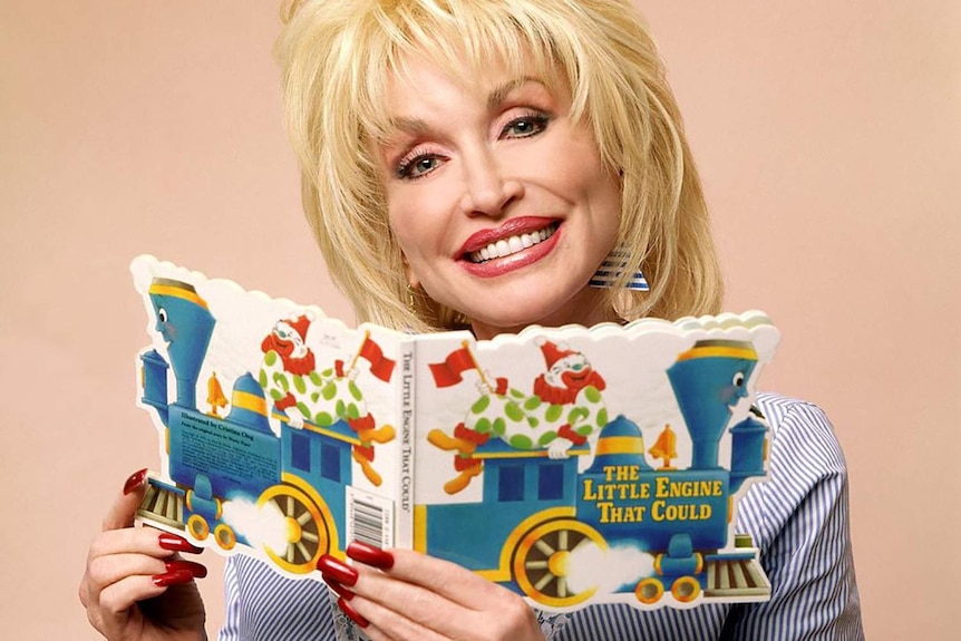 Dolly Parton smiles in a portrait, holding a picture book of 'The Little Engine that Could' up under her chin.