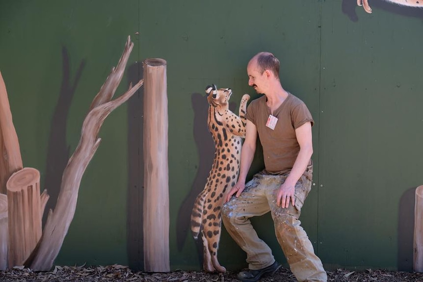 Man sit nexts to painting of a cheetah, posing as though the cheetah is jumping up on him.
