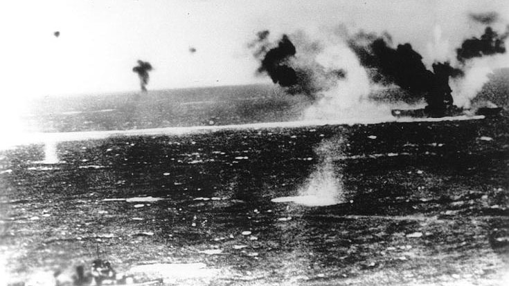 An old black and white photograph shows bombs being dropped on a warship in the sea.