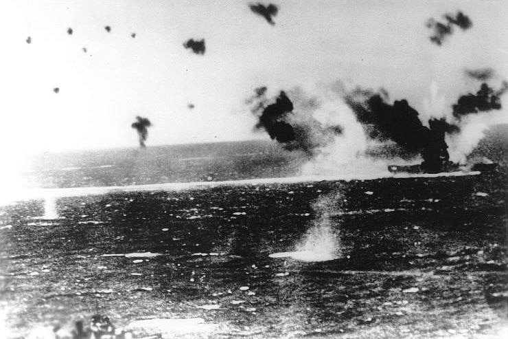 An old black and white photograph shows bombs being dropped on a warship in the sea.