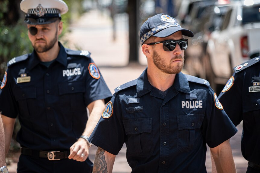 A man in NT Police uniform walking down the street, on a sunny day.