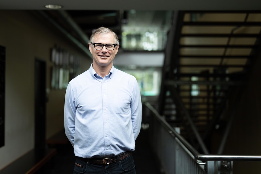 A man in a blue business shirt standing the hallways of the university. He has wispy grey hair and wears dark framed glasses.