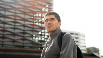 A man in a grey jumper with short hair and glasses looks into the distance with the sun coming off a grey and red building