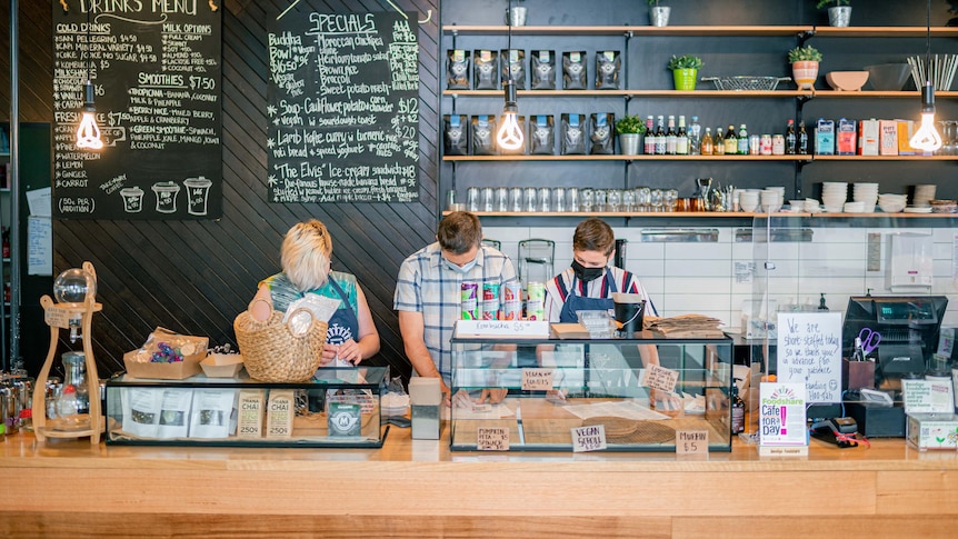 Three people standing behind the till and food cabinet at a cafe, wide shot with special board visible