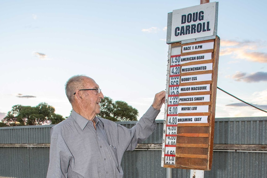An elderly man in a blue shirt, balding, wears glasses, stands next to a bookie board his names and number  slotted in.