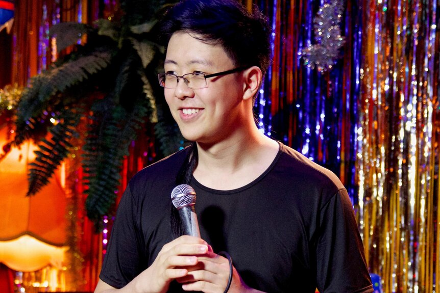 A man smiles and looks to the left of the camera as he holds a microphone in front of a sparkly tinsel wall.