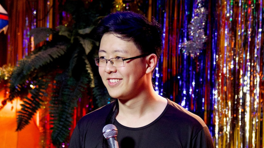 A man smiles and looks to the left of the camera as he holds a microphone in front of a sparkly tinsel wall.