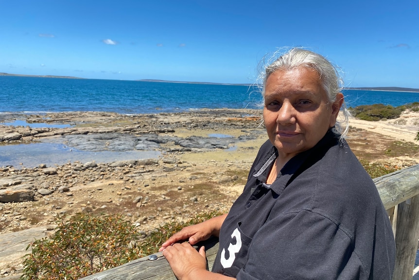 An Indigenous woman with long, silver hair stands in front of a rocky beach.