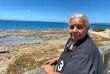 An Indigenous woman with long, silver hair stands in front of a rocky beach.