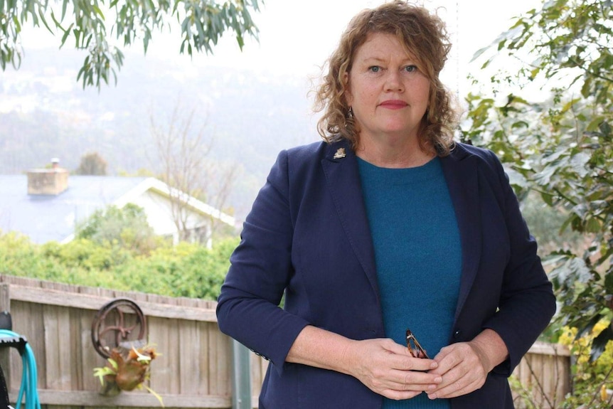 Anna Reynolds standing in her backyard. She has a straight face, curly fair hair and is wearing a navy jacket over a blue top.