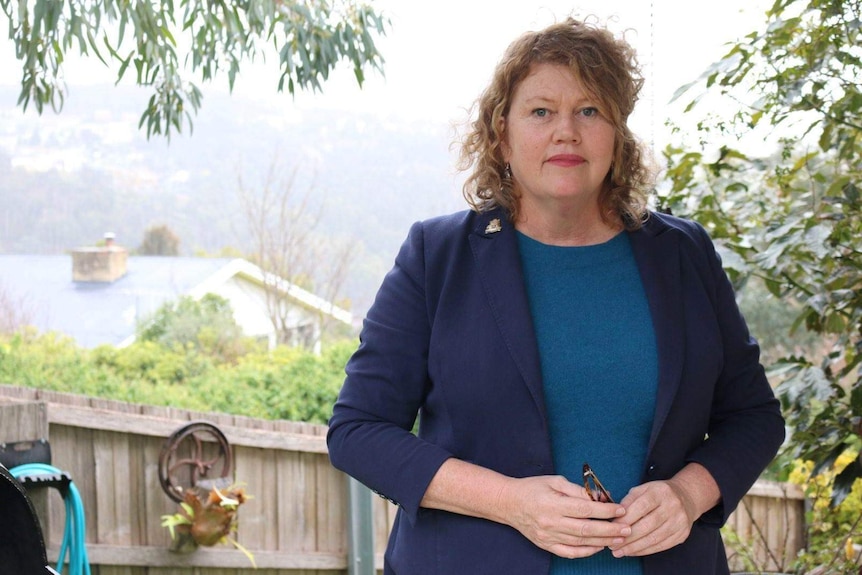 Anna Reynolds standing in her backyard. She has a straight face, curly fair hair and is wearing a navy jacket over a blue top.