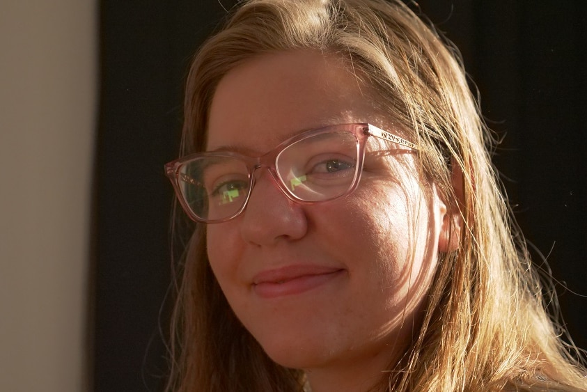 A young woman, Sarah Barclay, looks at the camera, with sunlight on her cheek.