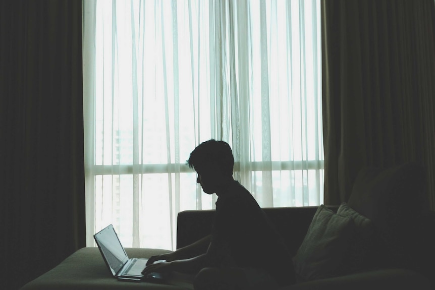 Man sitting on a bed in a room typing on his laptop