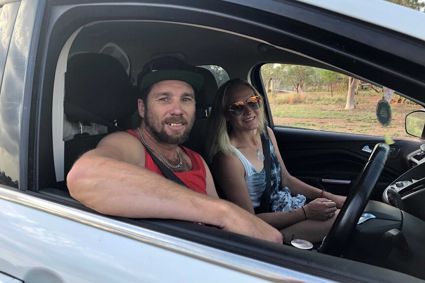 A man wearing a cap on his head sits in the driver's seat of a car next to a woman wearing sunglasses.