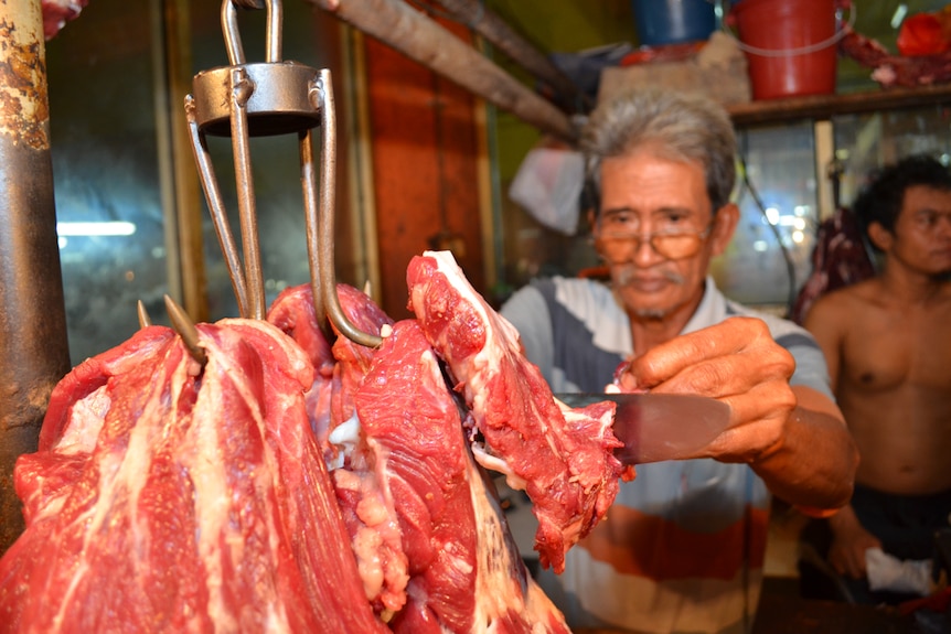 A man uses a large knife to slice at hunks of red meat hanging from hooks.