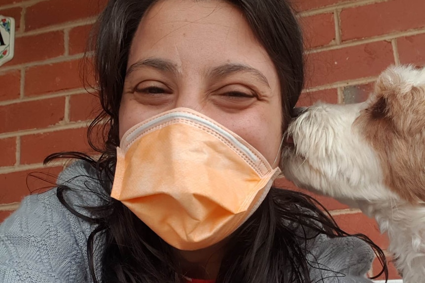 Close up of Laura, her dark hair around her shoulders and a surgical mask on. A brown and white dog sniffs her cheek. 