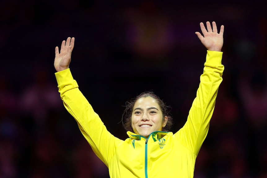 gymnast georgia godwin smiles and waves her hands in the air wearing a yellow jacket with australian coat of arms
