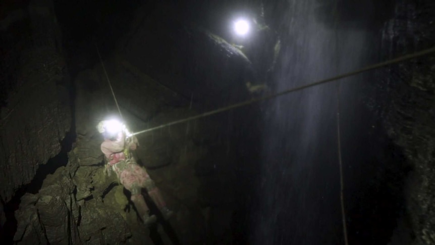 Man on rope in cave in front of waterfall.