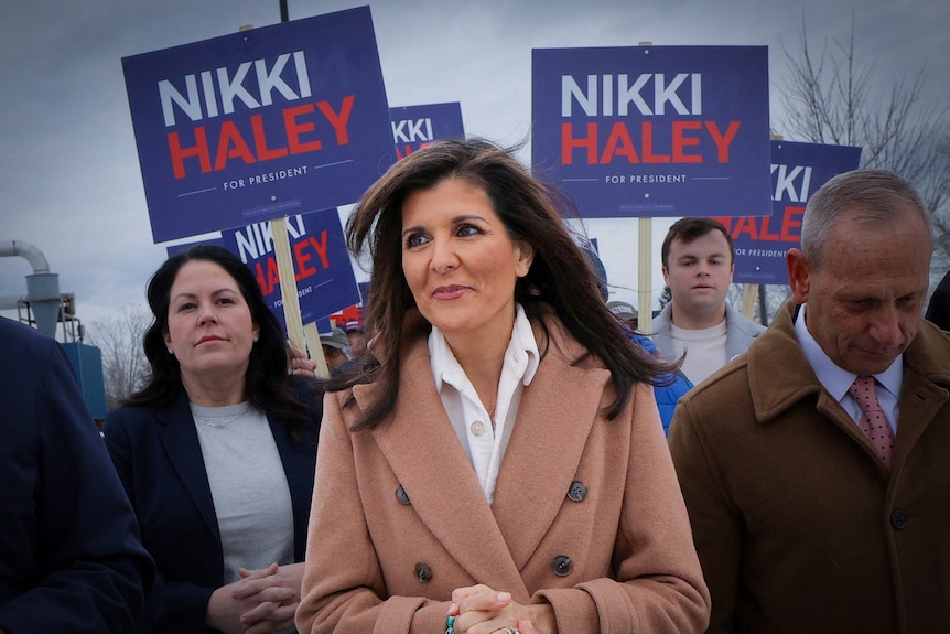 A woman in a camel coat walks through a crowd holding 'Nikki Haley' signs 