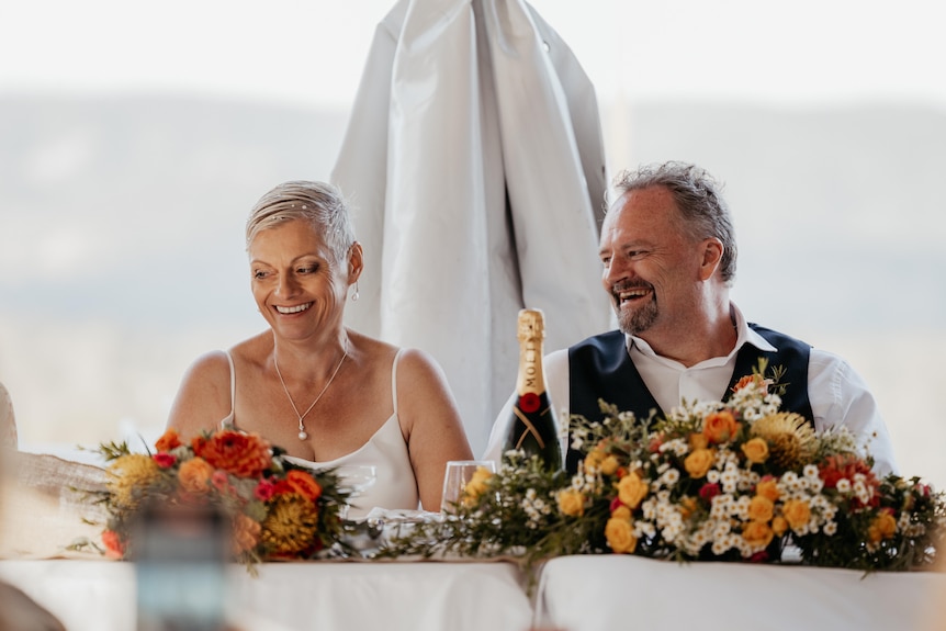 The married couple sit at the table smiling as they look over flowers and champagne. 