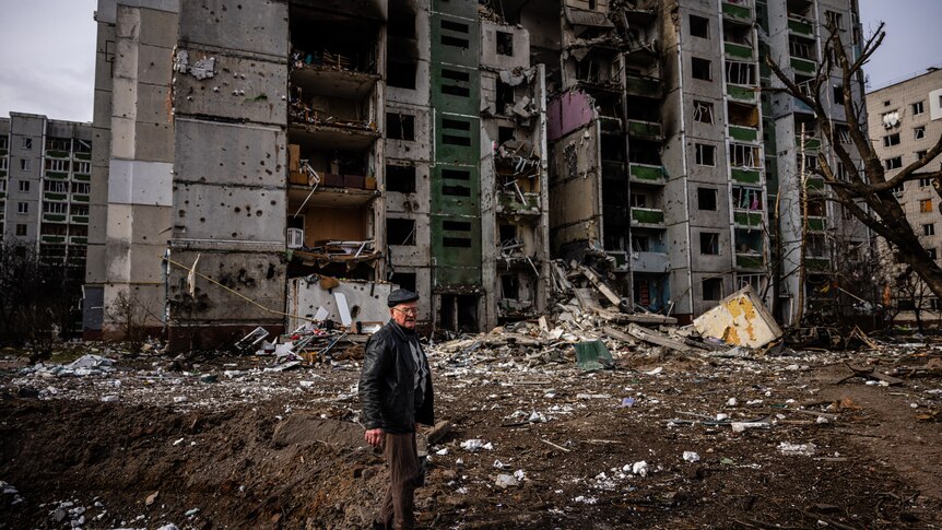 A man stands in front of a residential building damaged by shelling