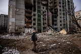 A man stands in front of a residential building damaged by shelling