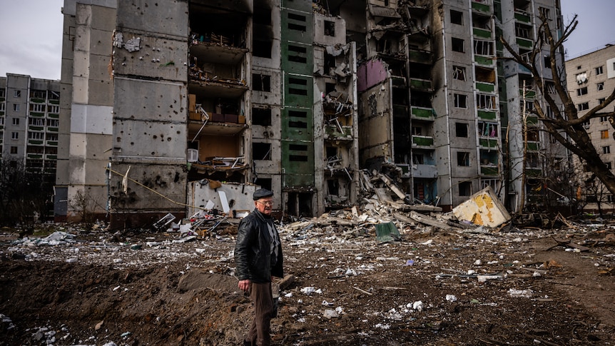 A man stands in front of a residential building damaged by shelling