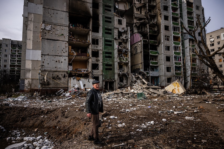 A man stands in front of a residential building damaged by shelling