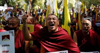 Monks wrapped in red robes hold flags and fists in the air, one man closes his eyes, yelling.
