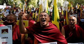 Monks wrapped in red robes hold flags and fists in the air, one man closes his eyes, yelling.