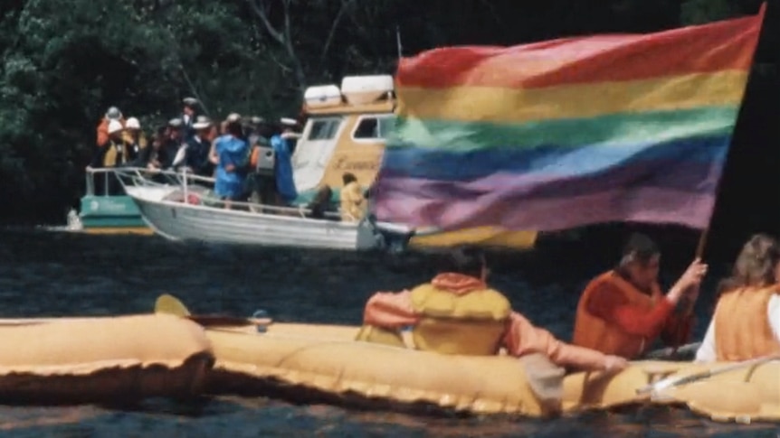 A man in a yellow rubber raft holding a rainbow flag.