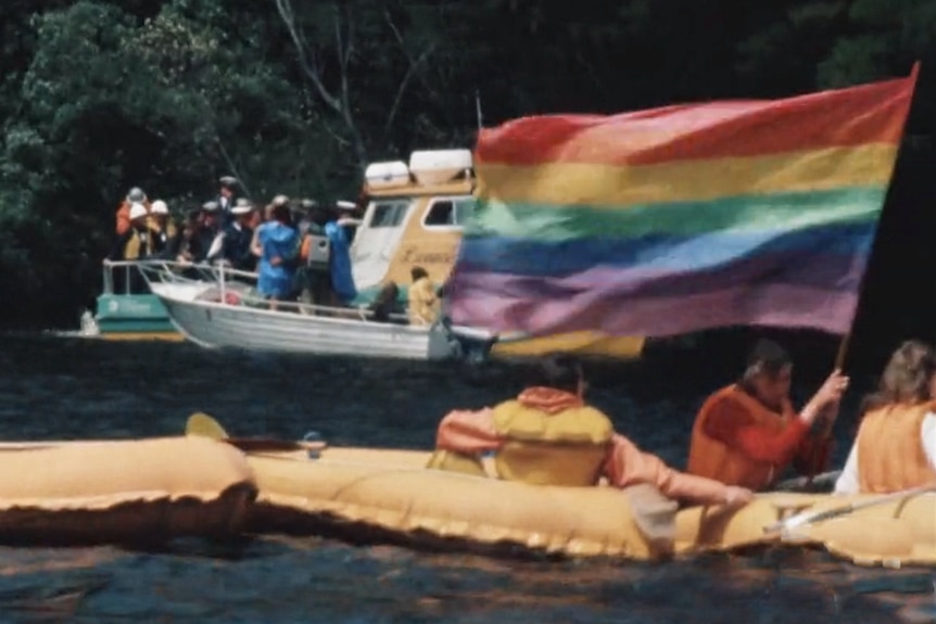 A man in a yellow rubber raft holding a rainbow flag.