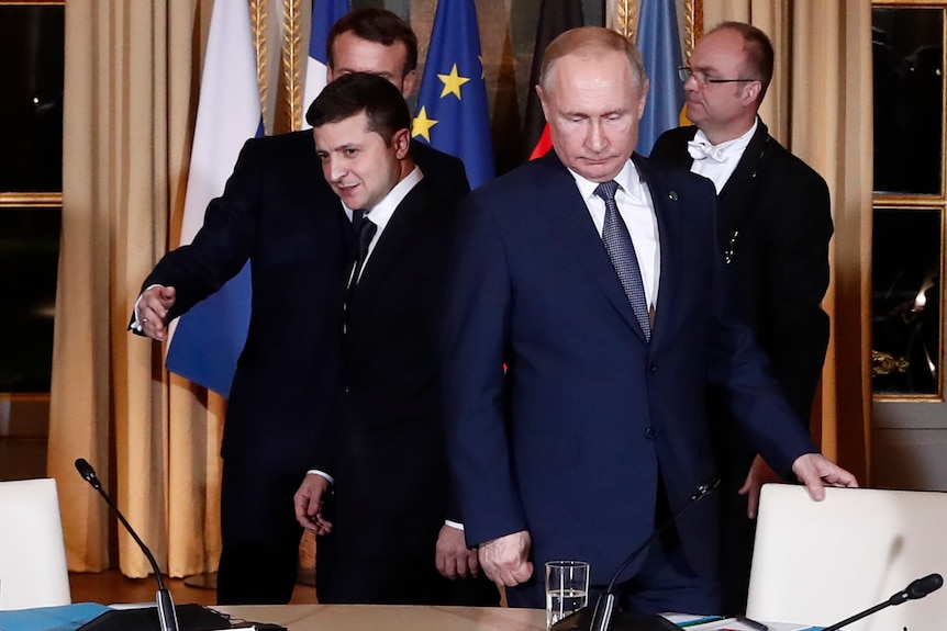 Volodymyr Zelenskyy and Vladimir Putin in suits approach their seats are two men stand behind as well as flags