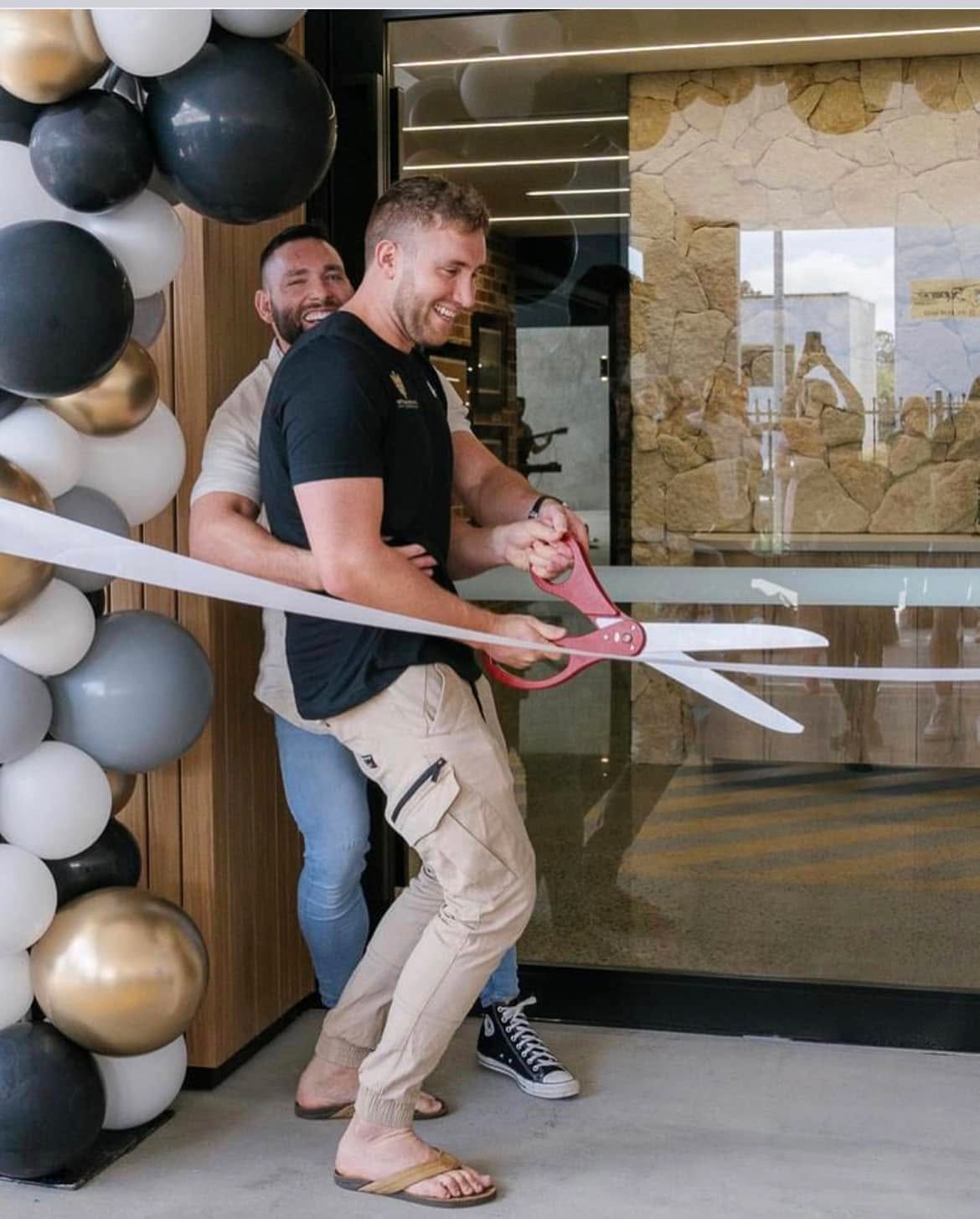 two men embracing each other while cutting ribbon with novelty-sized scissors