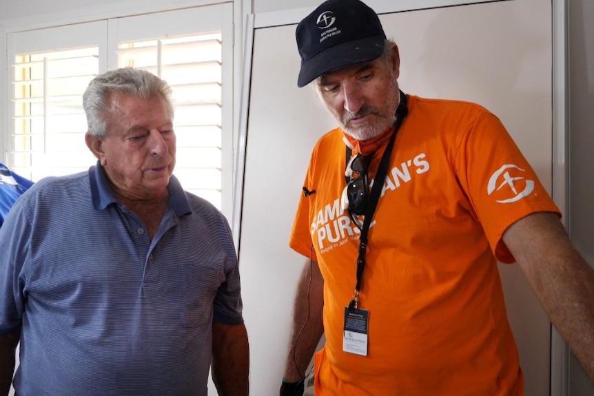 Two men stand looking at floor, inside a white room with slat blinds, younger man in orange volunteer T-shirt and cap 