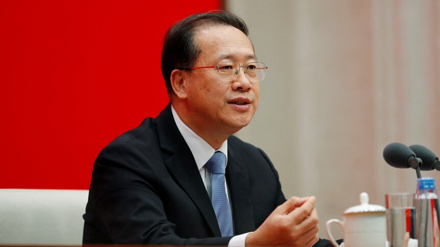 A Chinese man with thin-rimmed wire glasses gestures at table in front of microphones with bright red wall in background.