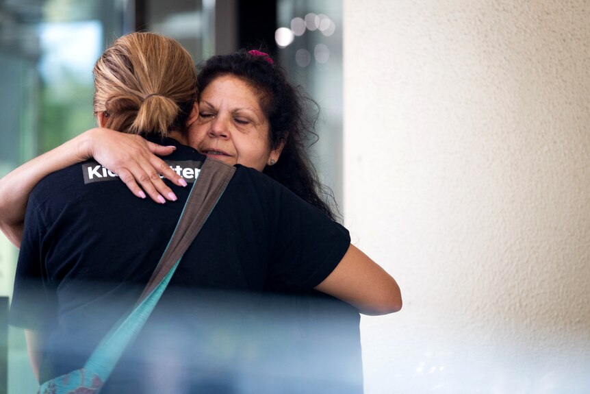 A woman hugs another woman outside of a white building
