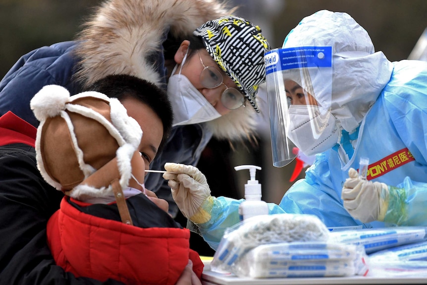 A medical staff in a protective suit takes a swab from a child as the child's parents watch.