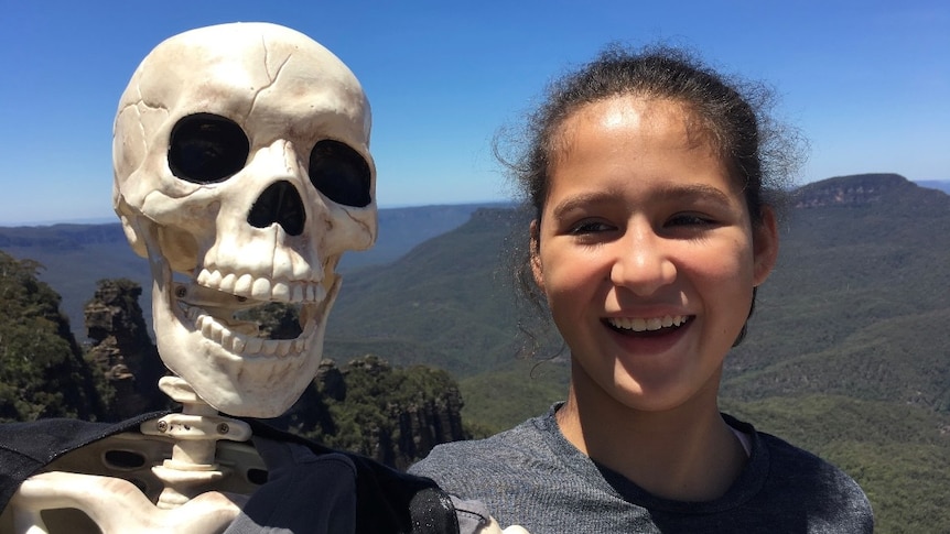 A young girl with dark hair smiles for a photo in front of a mountain range, alongside an adult-sized skeleton.