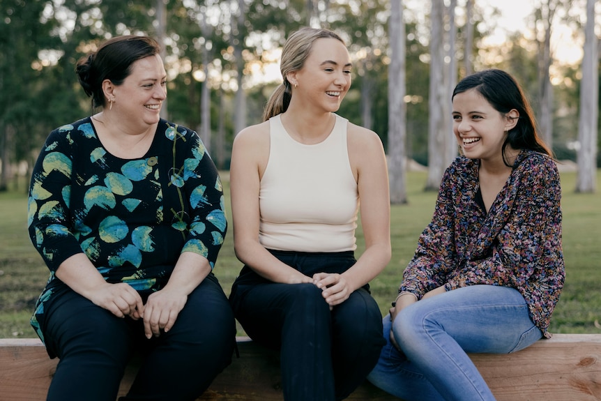 Woman in a blue and green top, a younger woman in a cream sleeveless top and a child in navy patterned shirt sit on park bench 