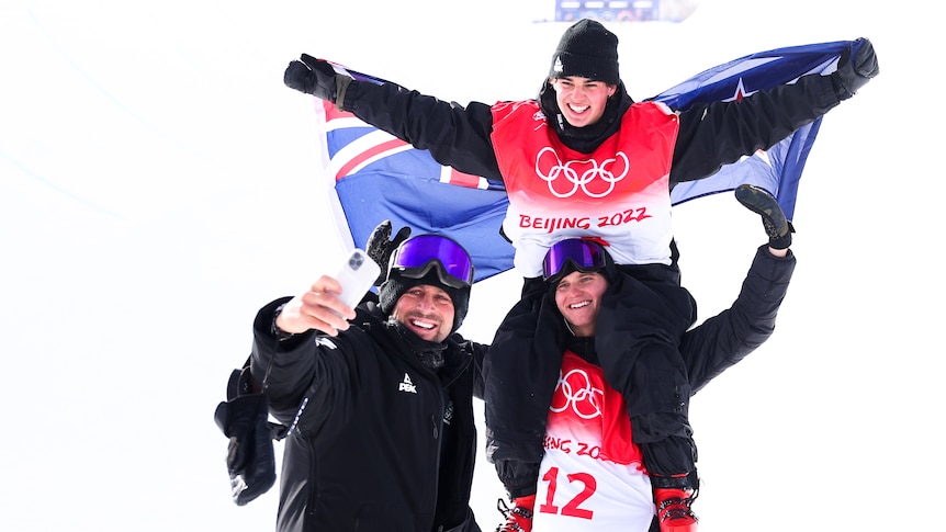 A man with a New Zealand flag sits atop another man's shoulders, as a third man takes a selfie