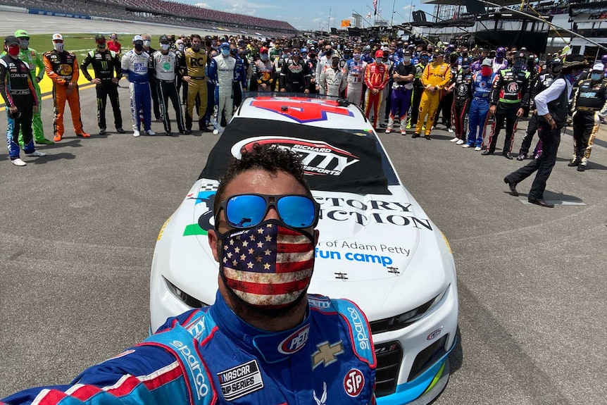 A man wearing dark glasses and a face mask stands in front of a race car with crews in background.