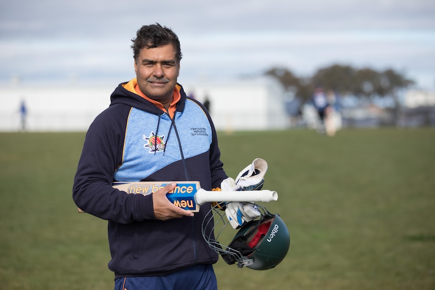 A man holding a cricket bat smiles at the camera.