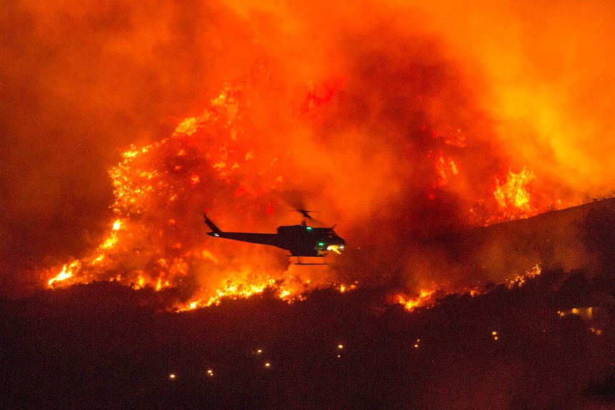 A helicopter hovers in the foreground of a fire tearing down the side of a mountain. The sky is orange.