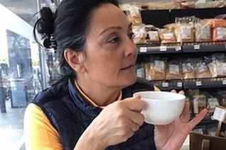 A dark-haired woman with her hair tied back is holding a coffee cup and looking to her right as she sits in a supermarket.