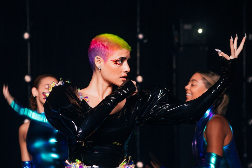 A woman wears a black pleather bodysuit that clashes with her technicolour hair. She sings into a microphone and holds a hand up