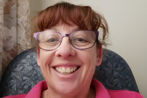 A woman wearing purple glasses and a pink work shirt sits in an office chair while smiling at the camera.