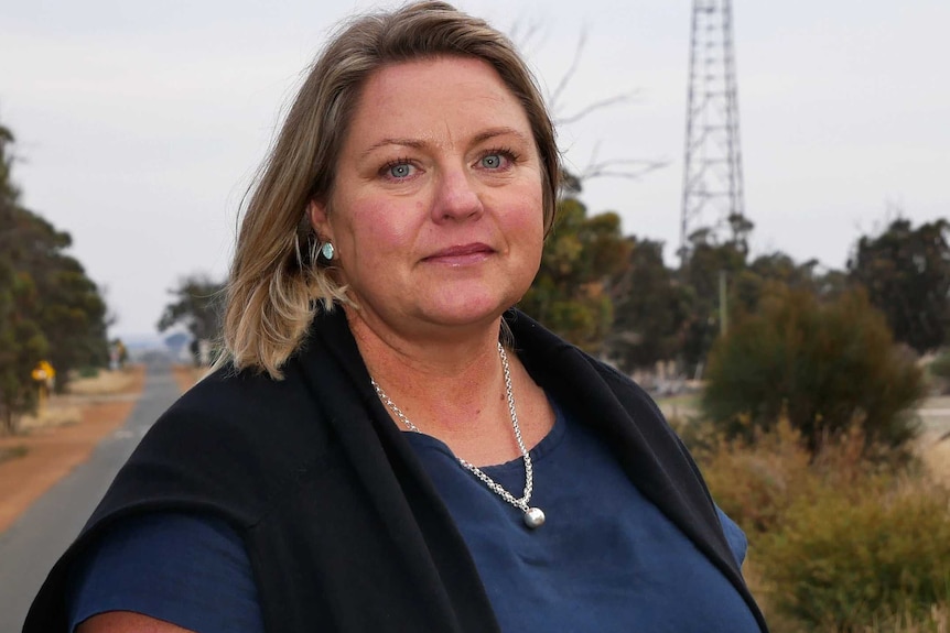 Jane Mary Richardson stands in front of a country road with a mobile phone tower in the dstance.