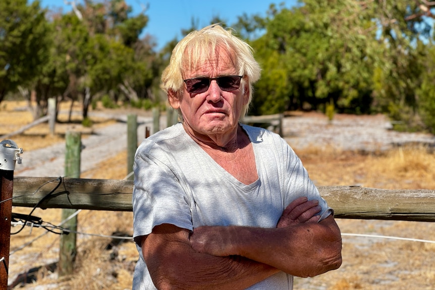 An older man in a white shirt and sunglasses stands on his bush property, looking stern